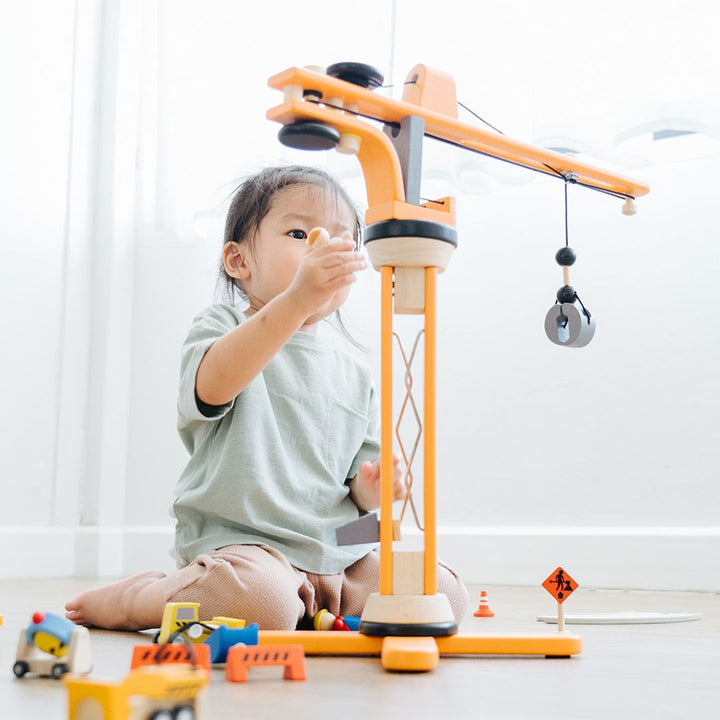 Child playing with the PlanToys Wooden Crane Construction Set, featuring a rotating crane with a working pulley, a concrete mixer, construction signs, barriers, and worker figurines.