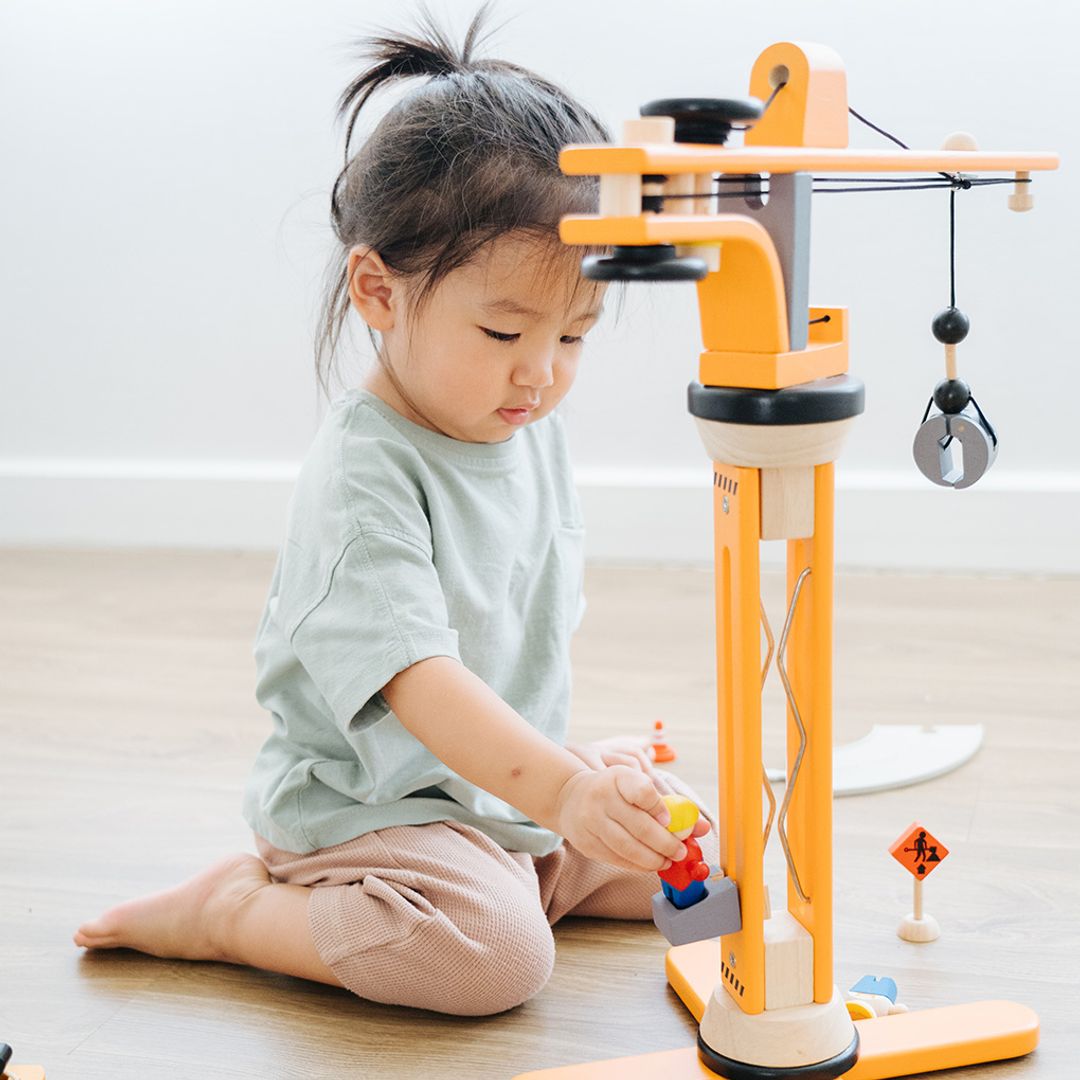 Child holding a wooden figure and looking down while playing the PlanToys Wooden Crane Construction Set, featuring a rotating crane with a working pulley, a concrete mixer, construction signs, barriers, and worker figurines.
