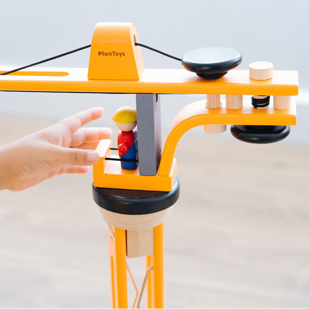 A close up of the top of the wooden PlanToys crane with a child's hand in front of one of the wooden figures who is in the tower.