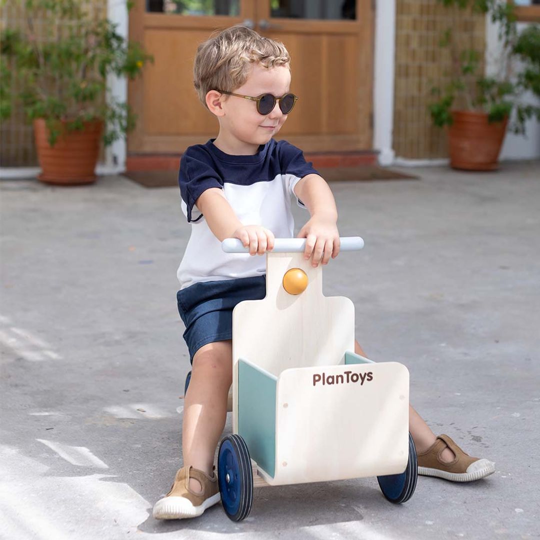 Child wearing sunglasses sitting on PlanToys Delivery Bike, a wooden ride-on toy with a front storage container holding the handlebars.