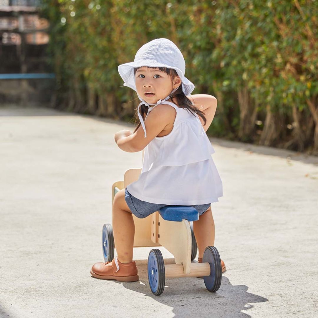 A oung child with a white sun hat sitting on the PlanToys Delivery Bike, a wooden ride-on toy with a front storage container, and the view is of her riding away down a driveway.