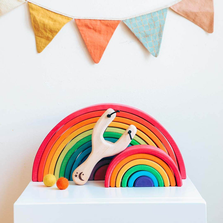 Colorful wooden rainbow toy with a slingshot on a white surface, with a colorful banner in the background.