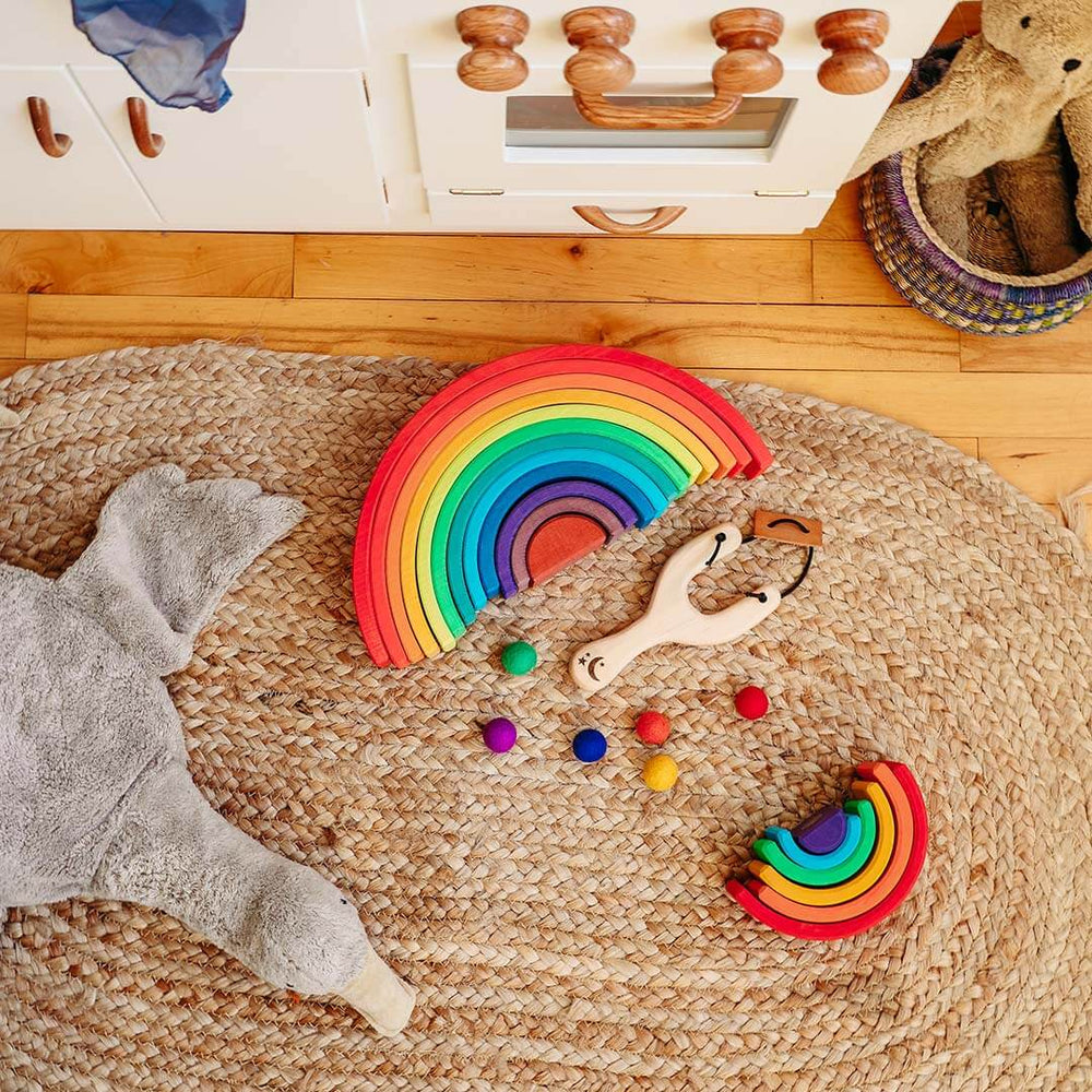 Colorful wooden rainbow toys on a woven mat with a wooden cabinet in the background.