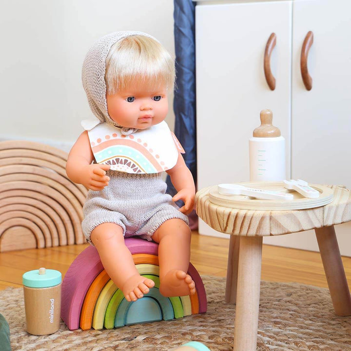 Toy doll with blonde hair sitting on a rainbow-colored toy in a room setting.