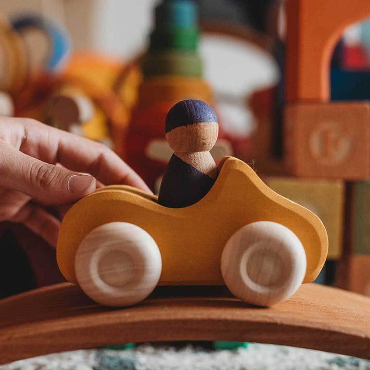 Wooden toy car with a small figure inside, held by a hand against a blurred background of toys.