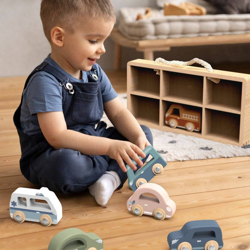 Child playing in playroom setting with the Speedy Monkey Wooden toy cars set with six colorful vehicles including a fire truck, ambulance, camper, and cars displayed in a wooden storage case - Bella Luna Toys