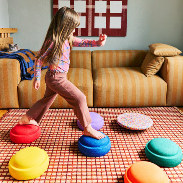 Young child stepping between a blue and red Stapelstein® stepping stones in a living room, with other rainbow-colored stones and a Super Confetti element arranged on a red-checked rug. A sofa and wall quilt are visible in the background.