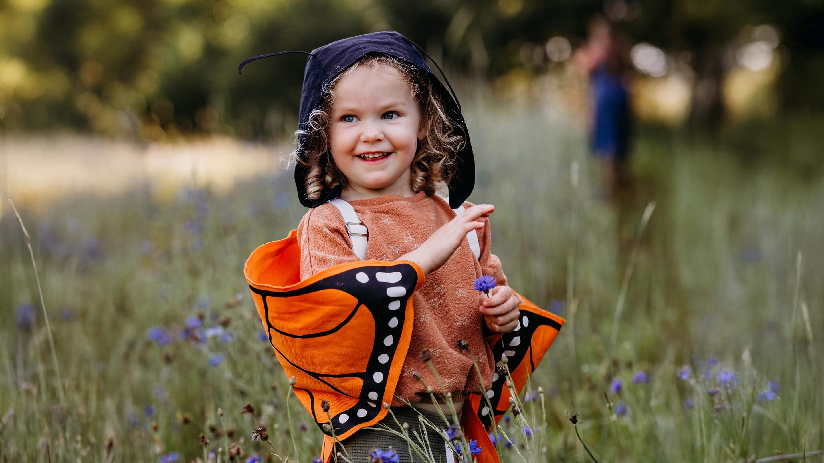 A young child wearing a butterfly costume stands in a grassy outdoor field. The costume includes an orange hood with black antennae, a brown top, and orange-and-black wing-like shoulder pieces.