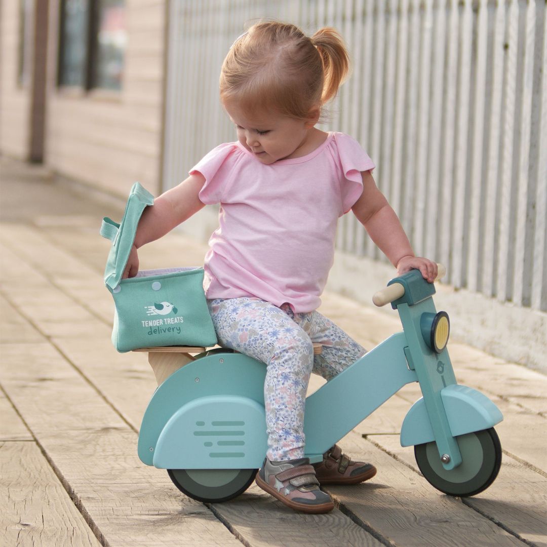 Child riding Tender Leaf Toys delivery scooter, placing items into the rear canvas delivery bag