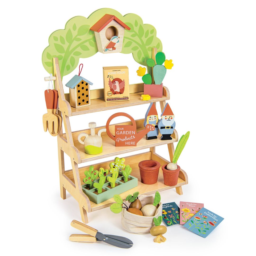 Wooden garden center play set from Tender Leaf Toys with three shelves displaying play pots, tools, seed packets, birdhouse, and two gnome figures. Includes miniature plants, gardening tools, and a watering can, all arranged on a wooden stand with a leafy arch.
