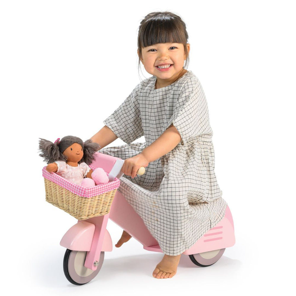 Young girl sitting on the Tender Leaf Toys pink scooter with a soft fabric doll in the wicker basket that is lines with pink gingham against a white background.